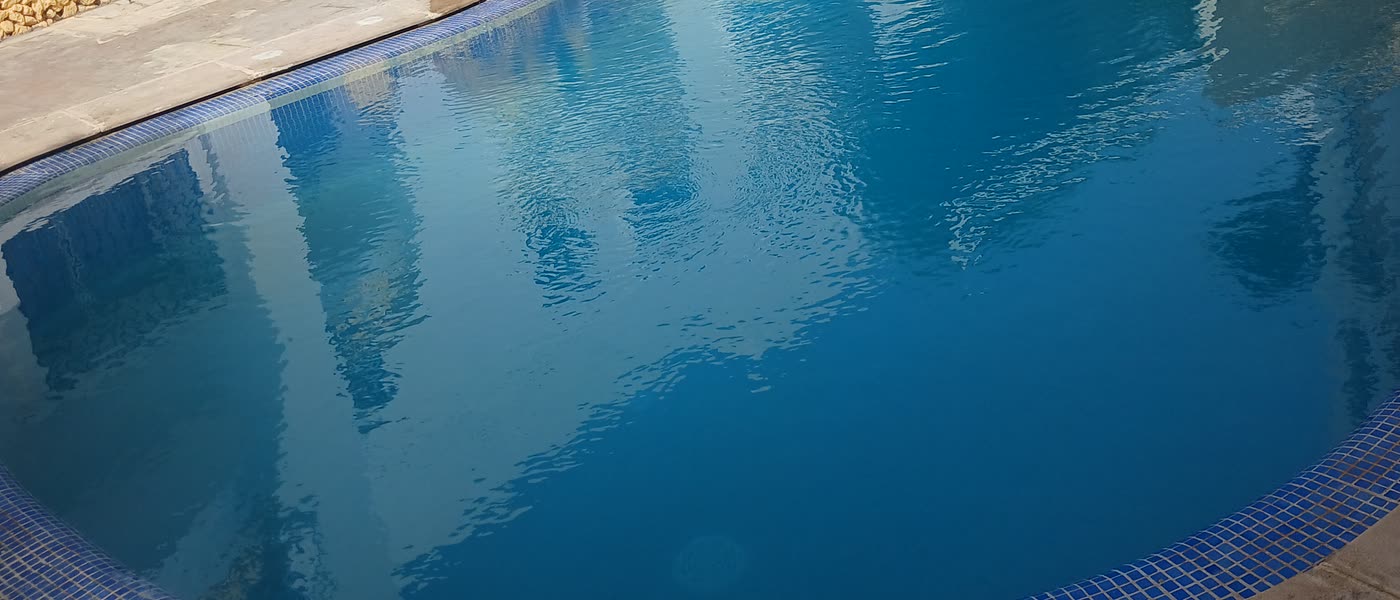 Gros plan sur la surface du grand bain : les façades blanches et leurs balcons s'y reflètent, la margelle en mosaïque bleue borde le bord.