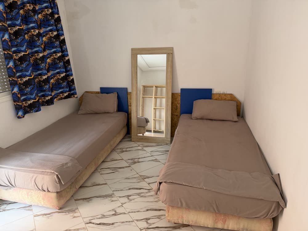 Chambre avec deux lits simples, têtes de lit bleues sur panneaux de bois, grand miroir posé au sol et rideau bleu à motif marbré.