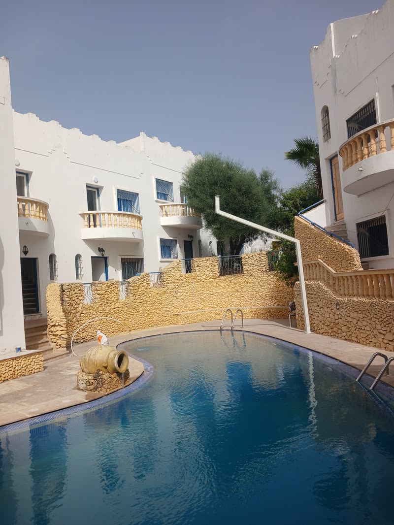 La piscine en plein soleil, l'arbre qui dépasse du mur de pierre ocre et les balcons blancs des duplex au fond.