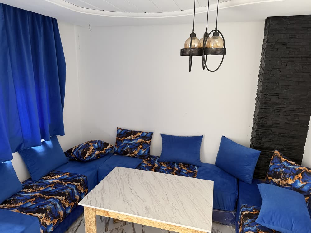 Salon marocain d'un duplex : banquettes bleues en U, coussins à motif marbré bleu et cuivre, table basse en marbre, mur en pierre noire et suspension à trois globes.